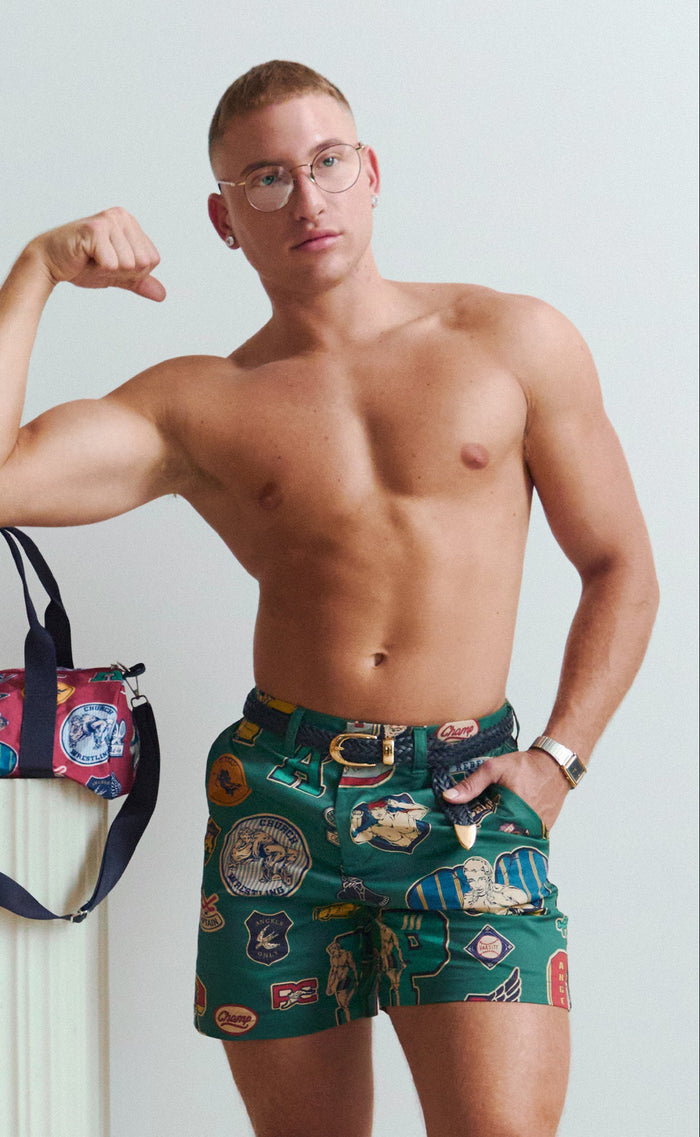A shirtless male model wearing green patterned swim trunks stands against a light gray studio background, flexing one arm. A colorful duffel bag rests on a white pedestal beside him.